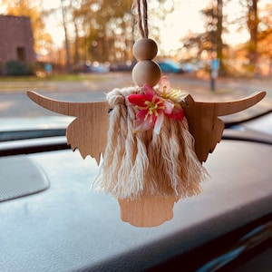 May include: A wooden cutout of a Highland cow head with a beige tassel fringe and a pink flower accent. The cow head is hanging from a rearview mirror in a car.