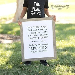 May include: A young boy wearing a black t-shirt with the words "The Plan" and two arrows printed on it. He is holding a white framed sign with the text "For 1,233 days, God was writing a chapter into my life called "FOSTER CARE" Today, He wrote "ADOPTED" August 05, 2020."