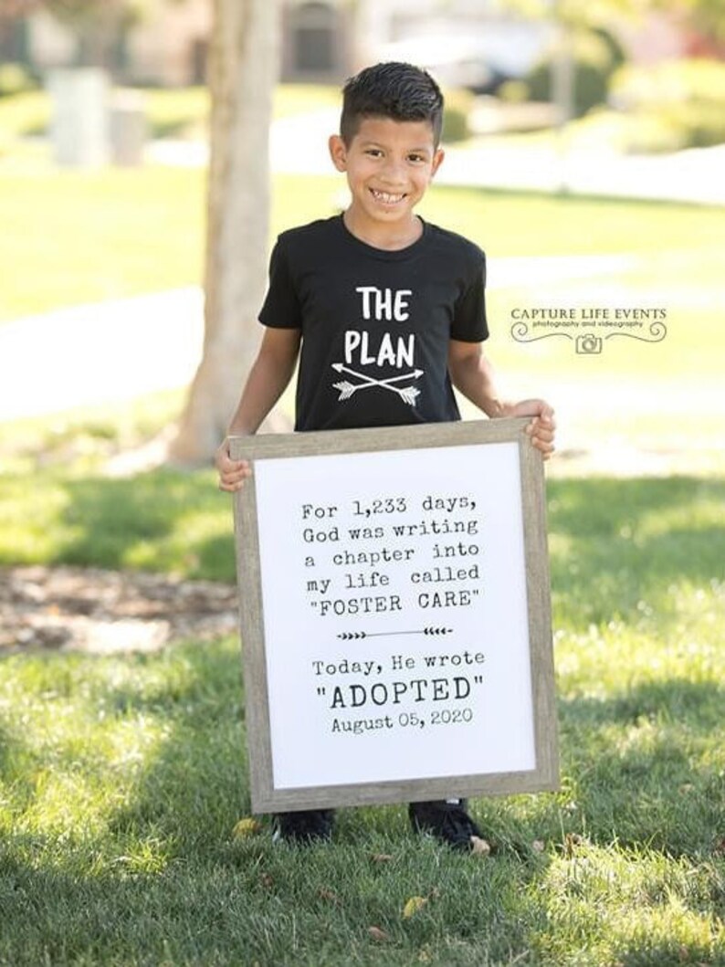 May include: A young boy wearing a black t-shirt with the words "The Plan" and two arrows printed on it. He is holding a white framed sign with the text "For 1,233 days, God was writing a chapter into my life called "FOSTER CARE" Today, He wrote "ADOPTED" August 05, 2020."
