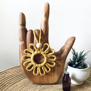 May include: A yellow macrame flower with a white wooden bead hangs from a wooden hand sculpture. A small potted succulent and a brown glass bottle are on a woven surface.