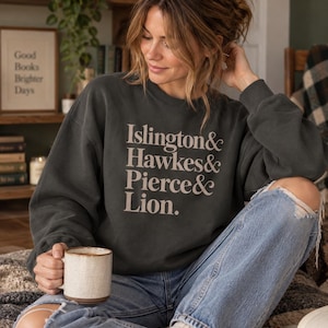 Puede incluir: Una sudadera de cuello redondo en color gris carbón oscuro con el texto "Islington & Hawkes & Pierce & Lion." impreso en una fuente serif de color beige claro en el pecho. La prenda tiene mangas largas y bordes acanalados en cuello, puños y dobladillo.