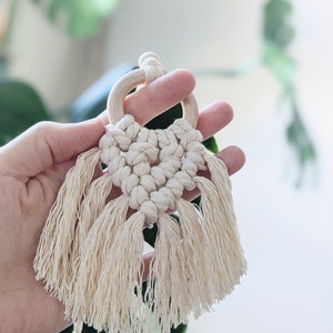Puede incluir: Un pequeño tapiz de macramé de color crema con un anillo de madera en la parte superior y largas borlas.