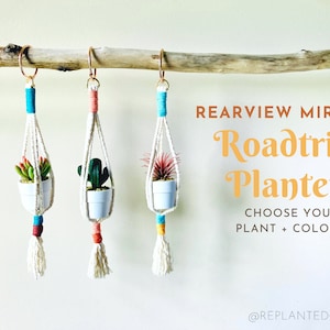 Puede incluir: Tres maceteros colgantes de macramé con pequeñas plantas en maceta colgando de una rama. Los maceteros están hechos con cuerda blanca y tienen detalles coloridos. El texto "Rearview Mirror Roadtrip Planter Choose Your Plant + Colors! @replantedgoods" es visible en la imagen.