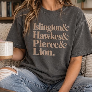 Pode incluir: Uma t-shirt de manga curta cinzento-carvão com o texto em tom bege Islington & Hawkes & Pierce & Lion. A peça tem gola redonda e uma textura de tecido desbotada de inspiração vintage, combinada com calças de ganga azul-claras.