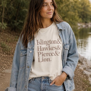 May include: A cream-colored short-sleeve t-shirt featuring the brown text Islington & Hawkes & Pierce & Lion. The shirt is worn under a light blue denim jacket and paired with matching blue denim jeans, creating a casual layered outfit.