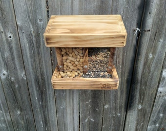 May include: A wooden bird feeder with two compartments, one filled with peanuts and the other with birdseed. The feeder has a clear plastic front to allow birds to see the food. The feeder is mounted on a wooden fence.