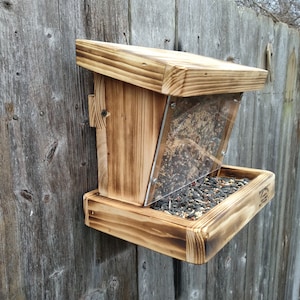 May include: A wooden bird feeder with a clear plastic front and a tray for seeds. The feeder is mounted on a wooden fence and is filled with birdseed.
