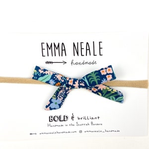 May include: A floral patterned headband with a blue background and pink, green, and yellow flowers. The headband is tied in a bow and is made by Emma Neale Handmade.