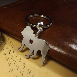 May include: Silver-toned key chain featuring a bulldog silhouette with a heart cutout. The keychain is attached to a silver-toned split ring.