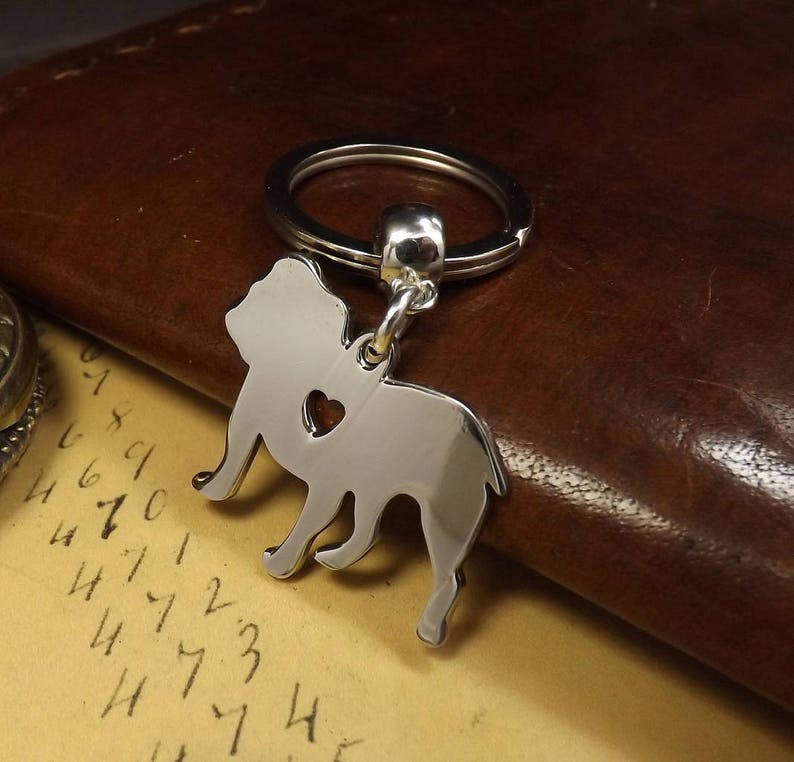May include: Silver-toned key chain featuring a bulldog silhouette with a heart cutout. The keychain is attached to a silver-toned split ring.