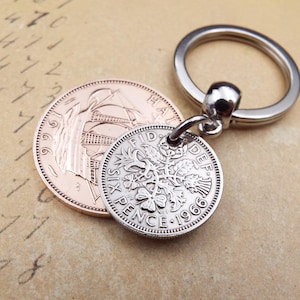 K&ouml;nnte beinhalten: Ein silberner Schl&uuml;sselanh&auml;nger mit einer 1966er Sixpence-M&uuml;nze mit floralen Details, befestigt an einem silbernen Schl&uuml;sselring. Eine kupferfarbene M&uuml;nze liegt neben dem Schl&uuml;sselanh&auml;nger. Der Hintergrund ist ein Vintage-Papier.
