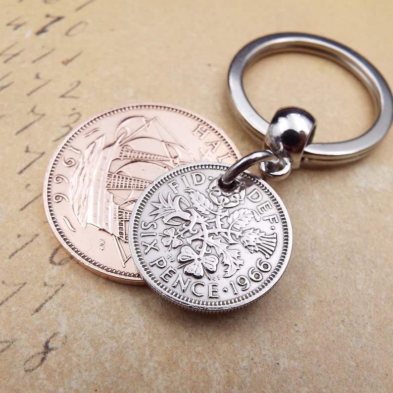 K&ouml;nnte beinhalten: Ein silberner Schl&uuml;sselanh&auml;nger mit einer 1966er Sixpence-M&uuml;nze mit floralen Details, befestigt an einem silbernen Schl&uuml;sselring. Eine kupferfarbene M&uuml;nze liegt neben dem Schl&uuml;sselanh&auml;nger. Der Hintergrund ist ein Vintage-Papier.