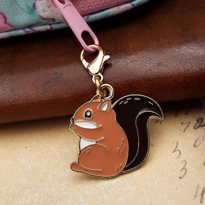 May include: A brown and black squirrel charm with a gold-coloured clasp and a pink zip pull. The squirrel has a white belly and a fluffy tail. The charm is attached to a brown leather surface.