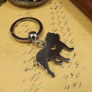 May include: Silver-toned keychain featuring a detailed silhouette of a dog with a heart cutout. The keychain has a silver-toned ring.