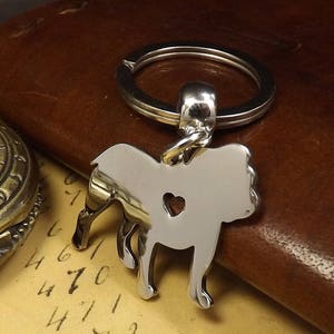 May include: Silver-toned keychain featuring a detailed silhouette of a bulldog with a heart cutout. The keychain is attached to a silver-toned ring.