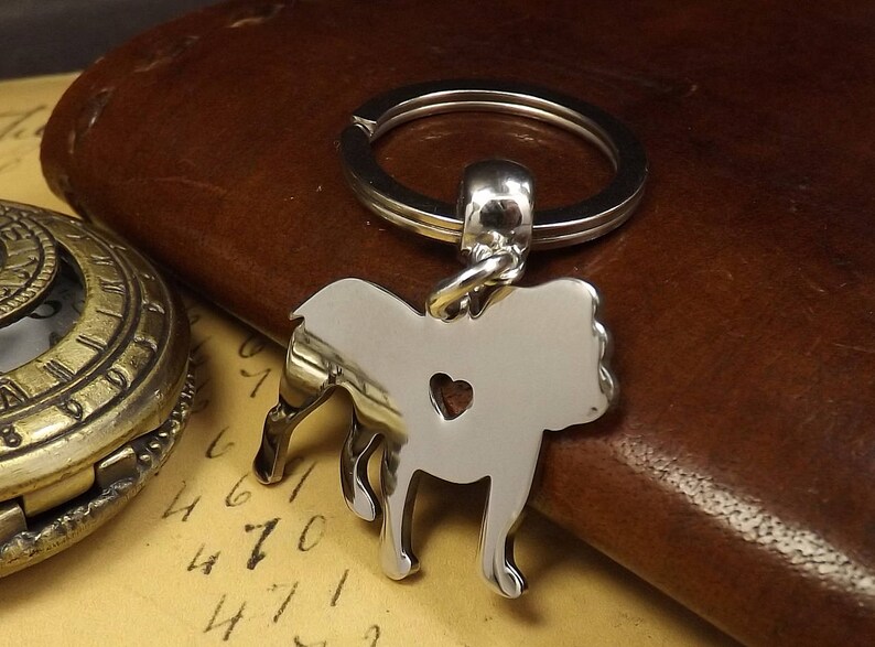 May include: Silver-toned keychain featuring a detailed silhouette of a bulldog with a heart cutout. The keychain is attached to a silver-toned ring.