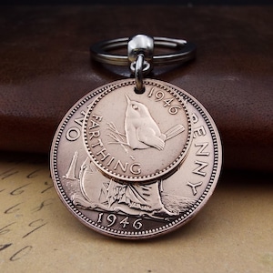 Pu&ograve; includere: Un portachiavi vintage in rame con due monete sovrapposte. La moneta superiore presenta un disegno di uccello e l'anno 1946. La moneta inferiore ha una figura femminile e la scritta "ONE FARTHING PENNY 1946".