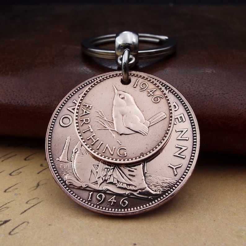 Pu&ograve; includere: Un portachiavi vintage in rame con due monete sovrapposte. La moneta superiore presenta un disegno di uccello e l'anno 1946. La moneta inferiore ha una figura femminile e la scritta "ONE FARTHING PENNY 1946".