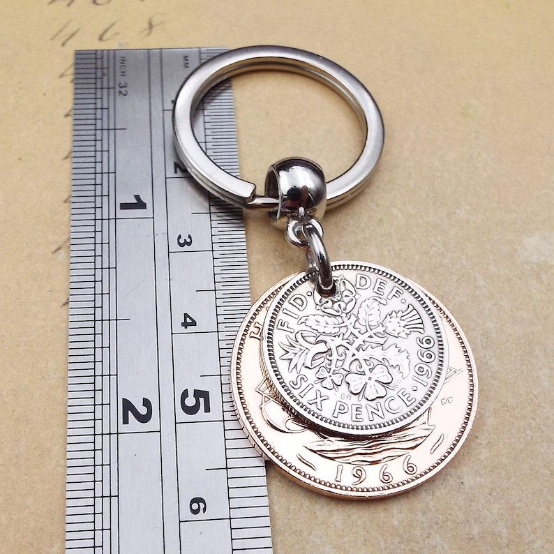K&ouml;nnte beinhalten: Ein silberner Schl&uuml;sselanh&auml;nger mit einem runden Ring und einer kleinen Kugel. Befestigt sind zwei antike M&uuml;nzen, eine silberne und eine kupferfarbene, mit dem Text "SIX PENCE" und dem Jahr "1966". Der Schl&uuml;sselanh&auml;nger ist etwa 5 cm lang.