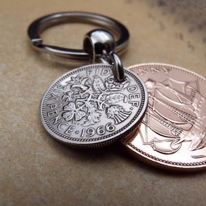K&ouml;nnte beinhalten: Ein silberner Schl&uuml;sselanh&auml;nger mit einer Six Pence M&uuml;nze von 1966 und einer kupferfarbenen M&uuml;nze. Die Six Pence M&uuml;nze zeigt florale Muster und die Aufschrift "Six Pence" und das Jahr "1966". Der Schl&uuml;sselanh&auml;nger ist an den M&uuml;nzen befestigt.