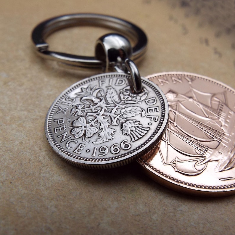 K&ouml;nnte beinhalten: Ein silberner Schl&uuml;sselanh&auml;nger mit einer Six Pence M&uuml;nze von 1966 und einer kupferfarbenen M&uuml;nze. Die Six Pence M&uuml;nze zeigt florale Muster und die Aufschrift "Six Pence" und das Jahr "1966". Der Schl&uuml;sselanh&auml;nger ist an den M&uuml;nzen befestigt.