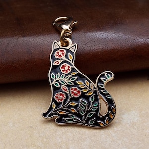 May include: A black cat charm with a gold-coloured clasp. The cat is decorated with red, yellow, and green floral designs. The charm is made of metal and has a glossy finish.