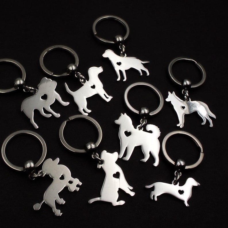 May include: A set of eight silver keychains featuring different dog breeds, each with a small heart cutout. The keychains are attached to silver key rings.