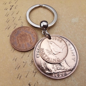 Puede incluir: Un llavero de color cobre con dos monedas antiguas y un anillo plateado. Una moneda es un penique, y la otra es una moneda de farthing de 1946, con un dise&ntilde;o de p&aacute;jaro. El fondo es una superficie beige con n&uacute;meros manuscritos.