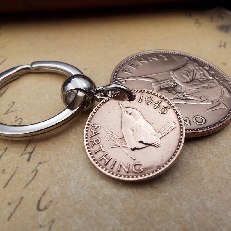 Puede incluir: Llavero con una moneda Farthing de 1946 con un dise&ntilde;o de p&aacute;jaro, sujeto a un llavero plateado. En el fondo se ve una moneda de Penny. Las monedas son de color cobre y est&aacute;n sobre un fondo neutro.