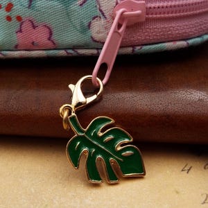 May include: A gold-coloured metal charm shaped like a green monstera leaf with gold trim. It's attached to a pink zip pull. The background features floral fabric and a brown leather surface.