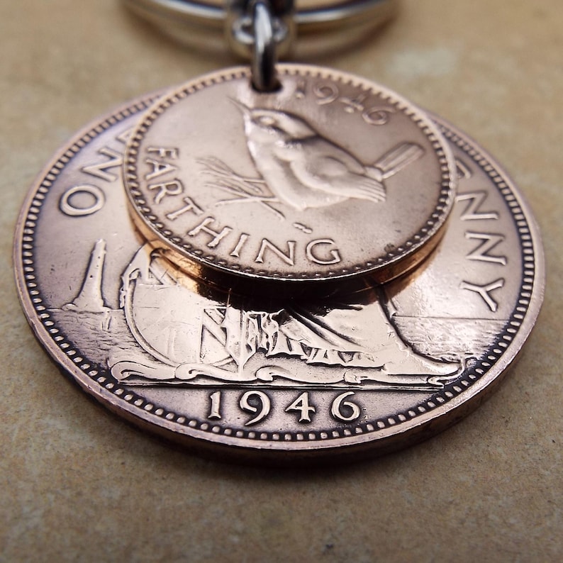 Puede incluir: Un colgante de monedas de cobre con dos monedas brit&aacute;nicas antiguas. La moneda m&aacute;s grande muestra el a&ntilde;o 1946 y una figura sentada, mientras que la moneda m&aacute;s peque&ntilde;a muestra un p&aacute;jaro y la palabra "FARTHING". El colgante est&aacute; unido a un anillo plateado.