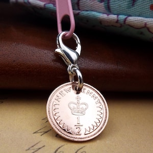 May include: A copper half penny coin charm with a silver clasp. The coin displays a crown and the words "NEW PENNY" and "1/2". The charm is attached to a pink zip, set against a brown leather background.