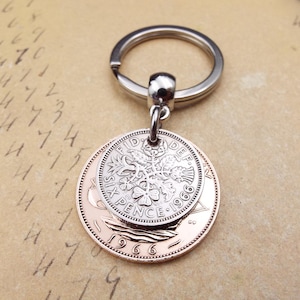 K&ouml;nnte beinhalten: Ein silberner Schl&uuml;sselanh&auml;nger mit einem Ring und einem Anh&auml;nger aus zwei M&uuml;nzen. Die obere M&uuml;nze ist silberfarben und tr&auml;gt die Aufschrift "SIX PENCE 1966". Die untere M&uuml;nze ist kupferfarben. Ein Vintage-Accessoire.