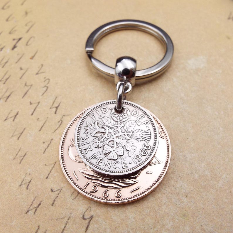 K&ouml;nnte beinhalten: Ein silberner Schl&uuml;sselanh&auml;nger mit einem Ring und einem Anh&auml;nger aus zwei M&uuml;nzen. Die obere M&uuml;nze ist silberfarben und tr&auml;gt die Aufschrift "SIX PENCE 1966". Die untere M&uuml;nze ist kupferfarben. Ein Vintage-Accessoire.