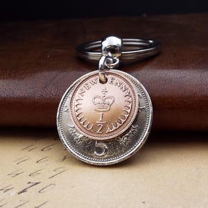 May include: A silver keyring featuring two coins. The top coin is copper and reads "NEW PENNY" around a crown. The bottom coin is silver and has the number "5". The keyring is attached to a silver ring.