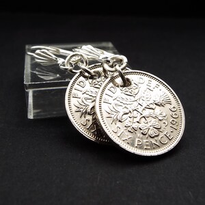 May include: A pair of silver coin earrings. Each earring is a round coin with embossed details, including text and floral designs. The earrings hang from silver lever-back closures. The coins appear vintage, with the year 1966 visible.