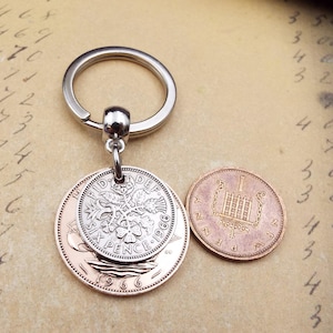 K&ouml;nnte beinhalten: Ein silberner Schl&uuml;sselanh&auml;nger mit einem Anh&auml;nger aus zwei alten M&uuml;nzen. Der Anh&auml;nger zeigt eine Six Pence M&uuml;nze von 1966 und eine kleinere M&uuml;nze. Die M&uuml;nzen sind an einem silbernen Ring befestigt.