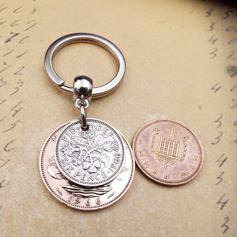 K&ouml;nnte beinhalten: Ein silberner Schl&uuml;sselanh&auml;nger mit einem Anh&auml;nger aus zwei alten M&uuml;nzen. Der Anh&auml;nger zeigt eine Six Pence M&uuml;nze von 1966 und eine kleinere M&uuml;nze. Die M&uuml;nzen sind an einem silbernen Ring befestigt.