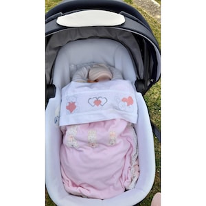 Peut inclure: Une couverture rose pour bébé avec des ours et des cœurs brodés est dans une poussette. La couverture est pliée sur le dessus de la poussette et le bébé est couché à l'intérieur.
