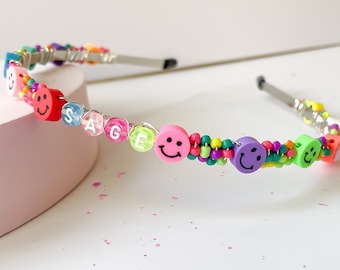 May include: A colorful beaded headband with the name "SAGE" spelled out in letter beads. The headband features smiley face beads in various colors, including red, pink, green, and purple, interspersed with smaller beads.
