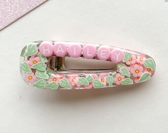 May include: A pink and green floral hair clip with the name "HAILEY" spelled out in pink letters.