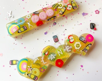 May include: Two yellow resin nameplates with glitter and colorful school-themed embellishments. The first nameplate is rectangular and says "ZOE". The second nameplate is shaped like a heart and says "LANE".