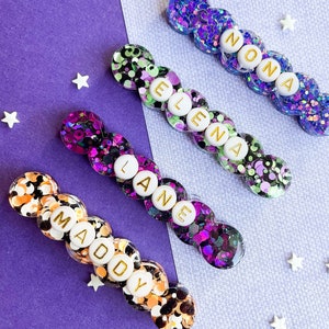 May include: Four glittery hair clips with names spelled out in gold letters. The clips feature a variety of colors, including purple, orange, and green, with the names "Maddy", "Lane", "Elena", and "Nona".