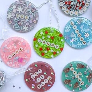 May include: A collection of seven circular resin ornaments featuring embedded colorful sprinkles, glitter, and white letter beads spelling names like Zoe, Danny, Lane, Winona, Keanu, and phrases like Merry Christmas and Let It Flow, each with a white string.