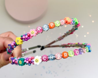 May include: Two colorful beaded headbands with the names "Poppy" and "Charlotte" spelled out in white letters. The beads are a mix of smiley faces, flowers, and solid colors.