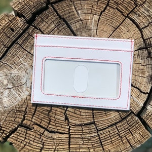 May include: White leather card holder with a red stitched border and a clear window for an ID card.