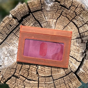 May include: A brown leather wallet with a red stitching detail and a clear window for an ID card.