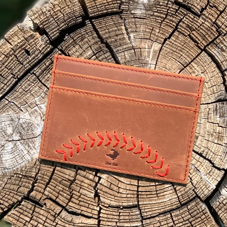 May include: A brown leather card holder with red stitching in the shape of a baseball. The card holder has the text "First Base" in white lettering.