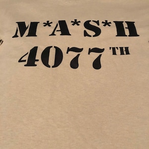 Könnte beinhalten: Ein beiges T-Shirt mit schwarzem Text, der "M*A*S*H 4077th" lautet.