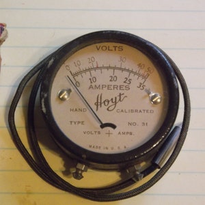May include: A black and white analog voltmeter with a white face and black lettering. The gauge reads "VOLTS" and "AMPERES" and is labeled "Hoyt". The gauge is calibrated to 50 volts and 35 amps. The gauge is made in the USA.